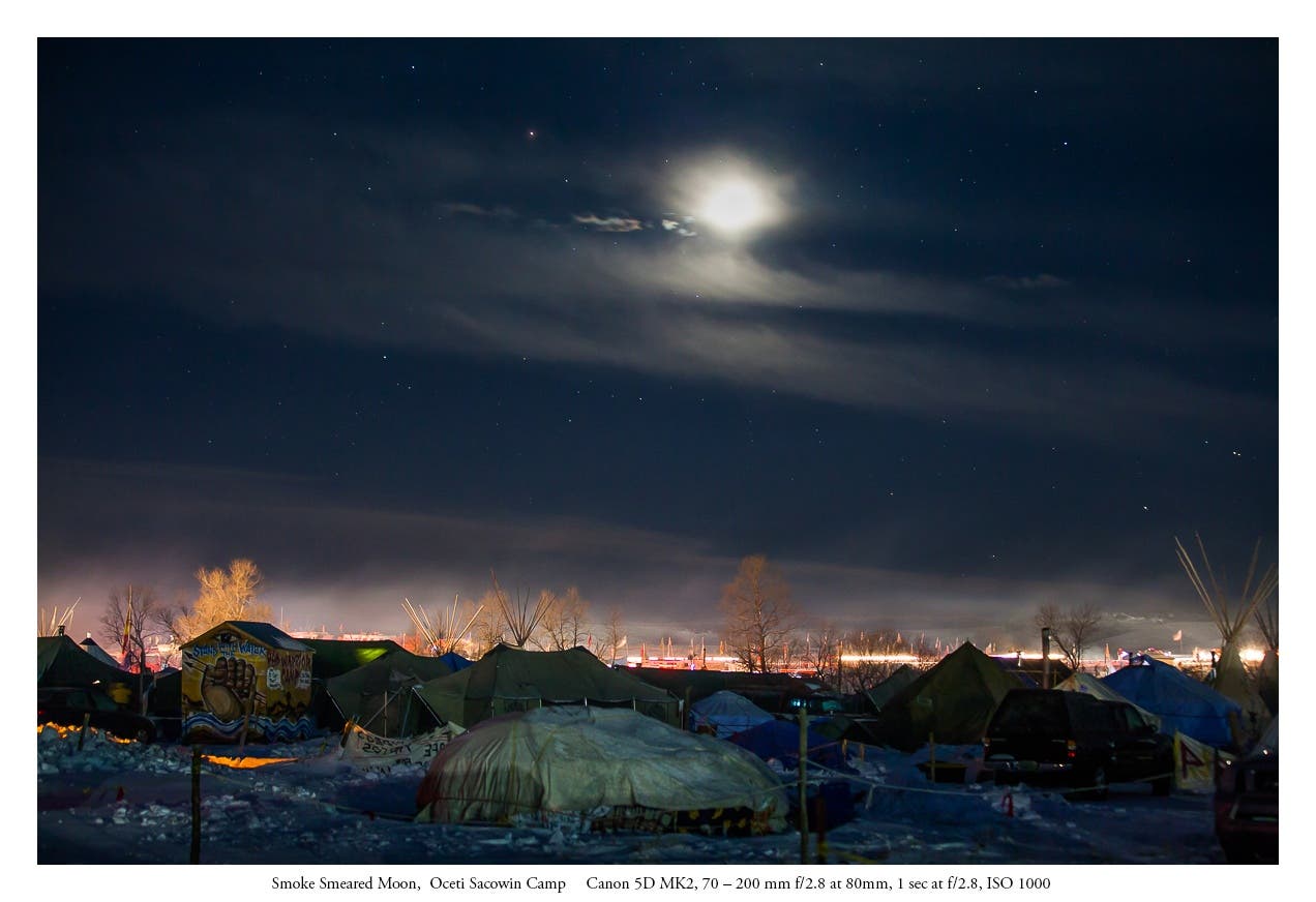 The ash rising from the Oceti Sacowin Camp reflects both the moon and the klieg lights.