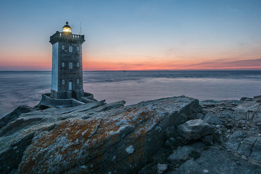 photo of lighthouse on rocky cliffs with pink sunset
