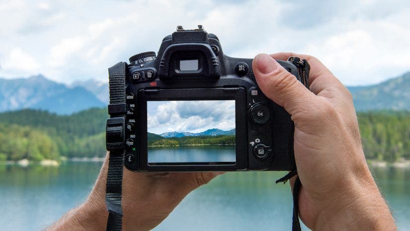 Hands of a male photographer holding a digital camera taking pictures of a idyllic landscape with a lake and mountains while the picture shows at the display