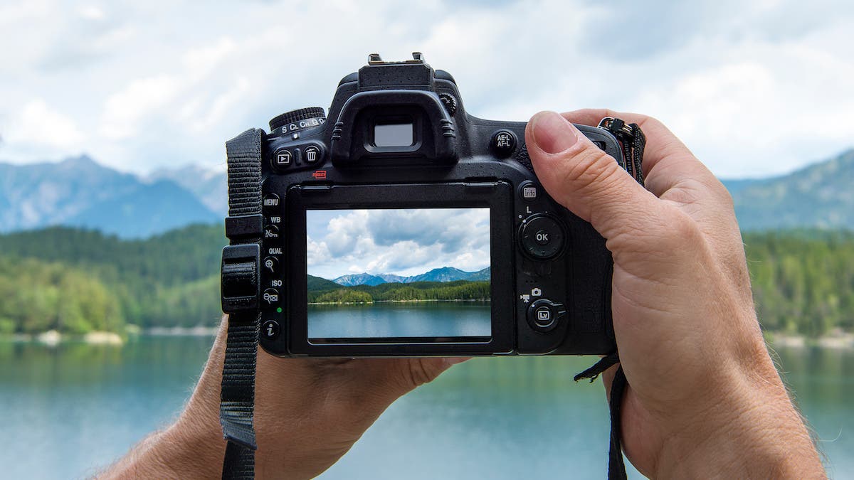 Hands of a male photographer holding a digital camera taking pictures of a idyllic landscape with a lake and mountains while the picture shows at the display