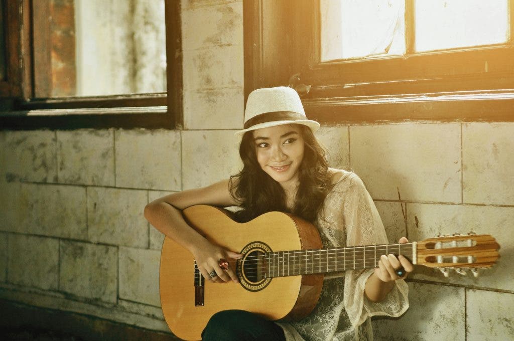 A girl holding a guitar while smiling and wearing a fedora