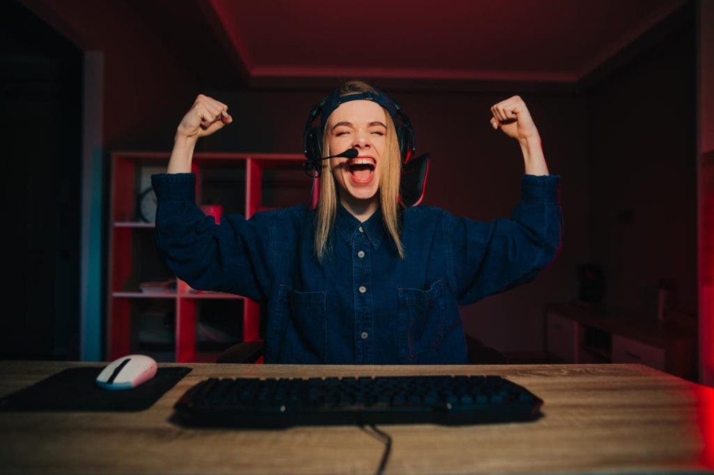 Happy female gamer in headset at night idol sits at the table and emotionally rejoices at the victory with raised hands.