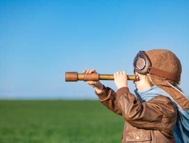 Happy child looking through spyglass outdoor in spring green field. Kid having fun against blue sky background. Imagination and freedom concept