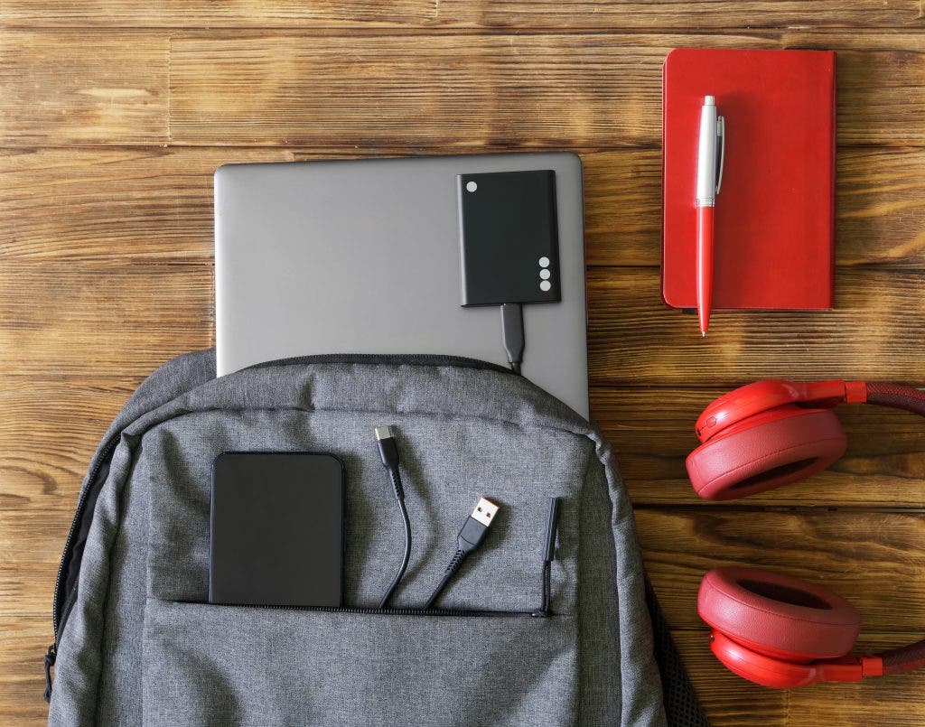 Gray textile urban backpack or bag with laptop, mobile phone and external ssd drive next to pen, notepad and headphones. Vintage wooden table. Accessories and gadgets for work and study. Close-up - Landscape photography gear.