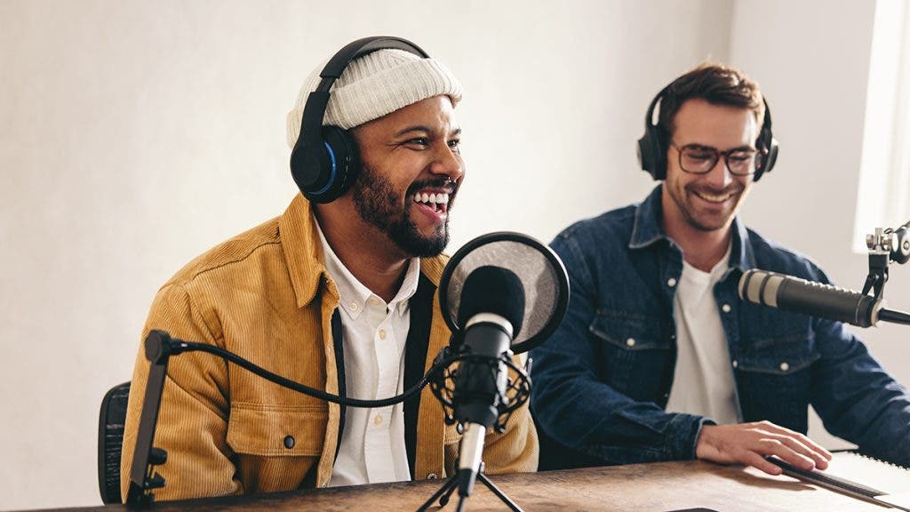 Happy radio presenter smiling while speaking into a microphone in a studio. Cheerful young man co-hosting an audio broadcast with a guest. Two young content creators recording an internet podcast.