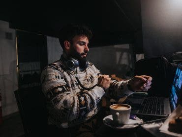 A concentrated male professional works on a laptop in a dimly lit room, with headphones around his neck and a cup of coffee nearby.