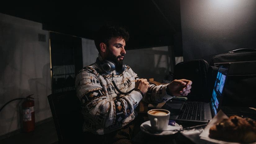 A concentrated male professional works on a laptop in a dimly lit room, with headphones around his neck and a cup of coffee nearby.