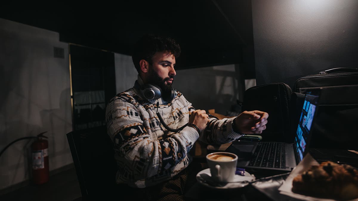 A concentrated male professional works on a laptop in a dimly lit room, with headphones around his neck and a cup of coffee nearby.