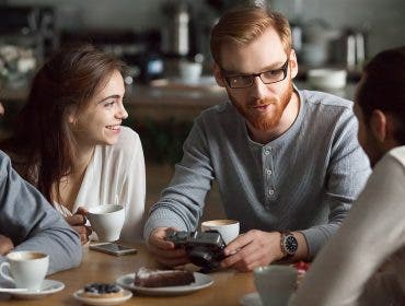 Young redhead guy holding camera showing photos sharing recent memories or travel impression hanging with diverse friends, smiling multiracial millennial people talking sitting together at cafe table