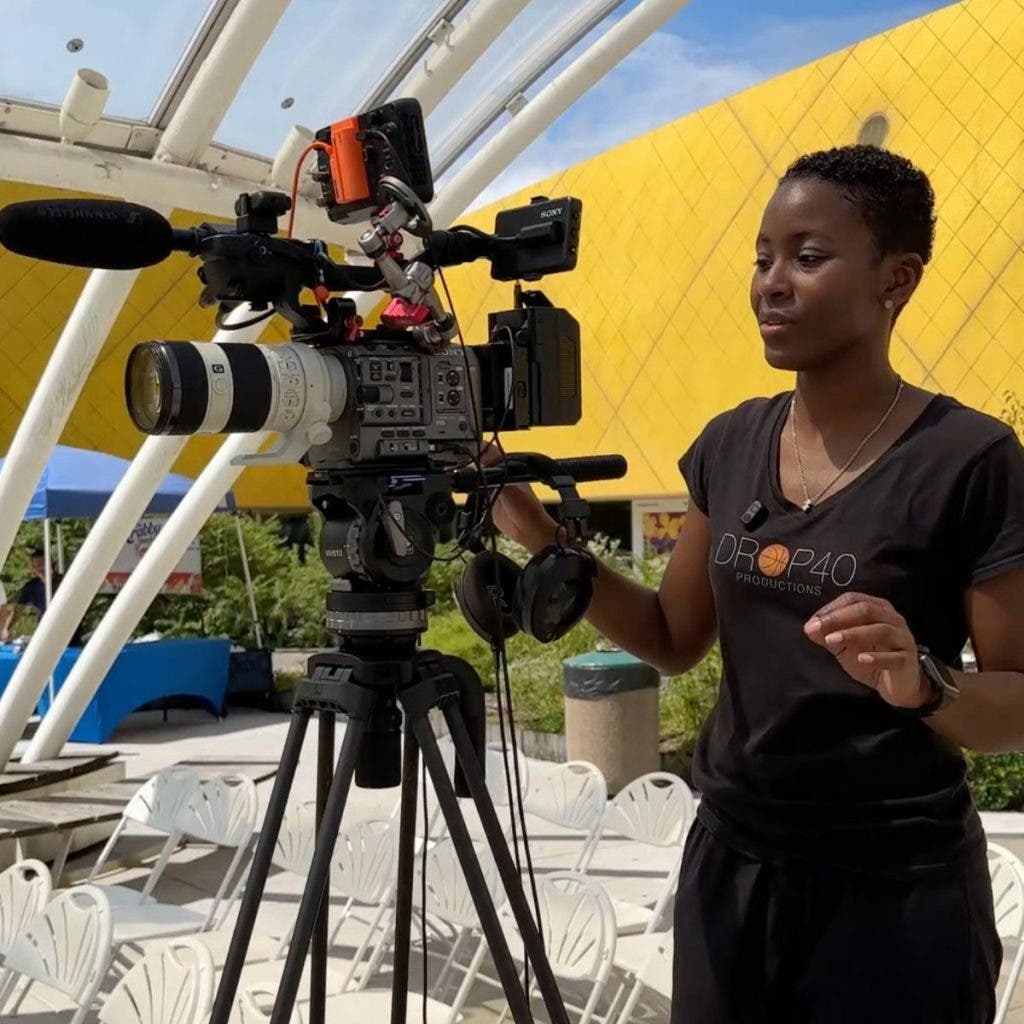 A videographer stands behind a rigged Sony FX6 on a tripod, operating the main camera for a two-camera setup at an outdoor event.