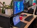 A comprehensive gaming workstation featuring a dual-monitor setup with a laptop serving as a secondary screen, an external mechanical keyboard, a red controller, and a large battery backup unit.
