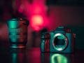 A professional mirrorless camera body and a high-end lens standing on a dark surface, illuminated by moody pink and teal studio lighting to illustrate the concept that the right gear pays for itself.