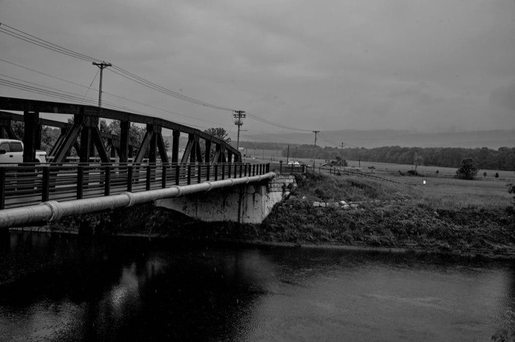 A wide-angle black and white landscape featuring a metal truss bridge crossing a dark river under an overcast, moody sky. Shot by Daniel Norton on a Leica M11 Monochrom.