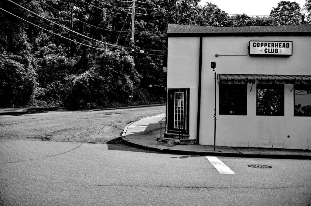 A high-contrast black and white street photograph of a white corner building featuring a sign that reads "COPPERHEAD CLUB." Power lines crisscross the sky above a dark, wooded area in the background. Shot by Daniel Norton on a Leica M11 Monochrom.
