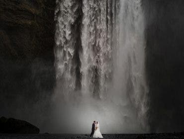 A wedding couple standing at the base of a massive, powerful waterfall, highlighting the epic scale of the landscape, shot by Susan Stripling.