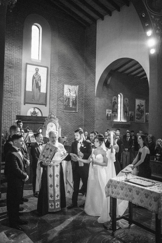 A black and white capture of a traditional religious wedding ceremony inside a grand brick-walled church, showing the priest with a liturgical book and the couple holding candles; essential coverage for a wedding photography side hustle. Photo by Susan Stripling.