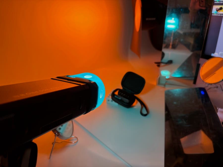 A close-up of a studio lighting setup showing a flash strobe equipped with a glowing blue gel modifier aimed at a small black case on a white table. The background is bathed in a bright orange light, and a small makeup mirror is used to reflect the light onto the product.
